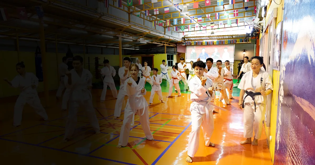 บรรยากาศการฝึกในโถงฝึกของ IMAC Dojo ผู้เรียนหลายวัยยืนตั้งการ์ดเป็นแถว มีหุ่นไม้และกระสอบอยู่ด้านหลัง